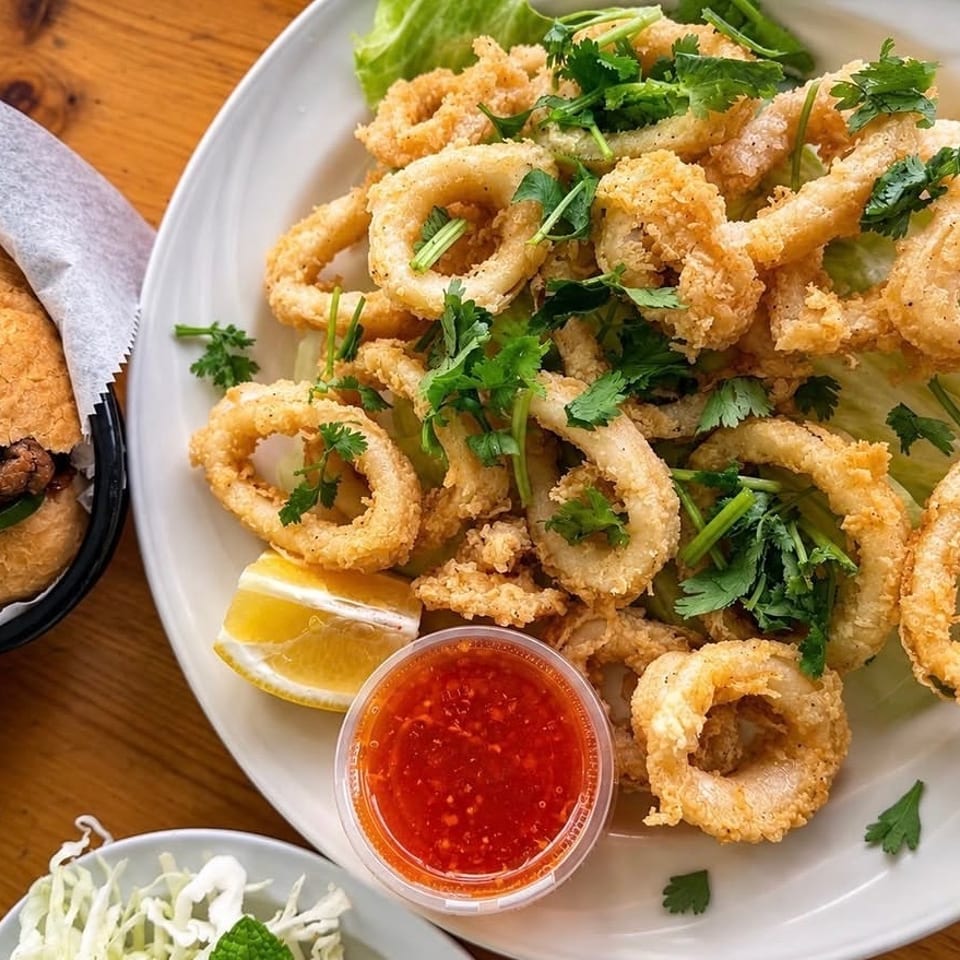 Crispy fried calamari rings garnished with cilantro, served with lemon wedges and sweet chili sauce beside a sandwich.