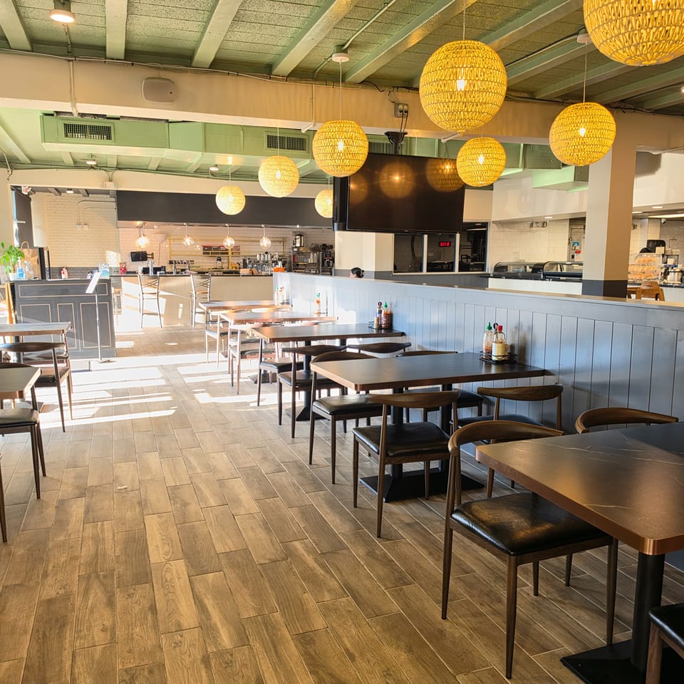 Bright modern restaurant dining room with wooden tables, black chairs, woven pendant lights, and an open kitchen counter.