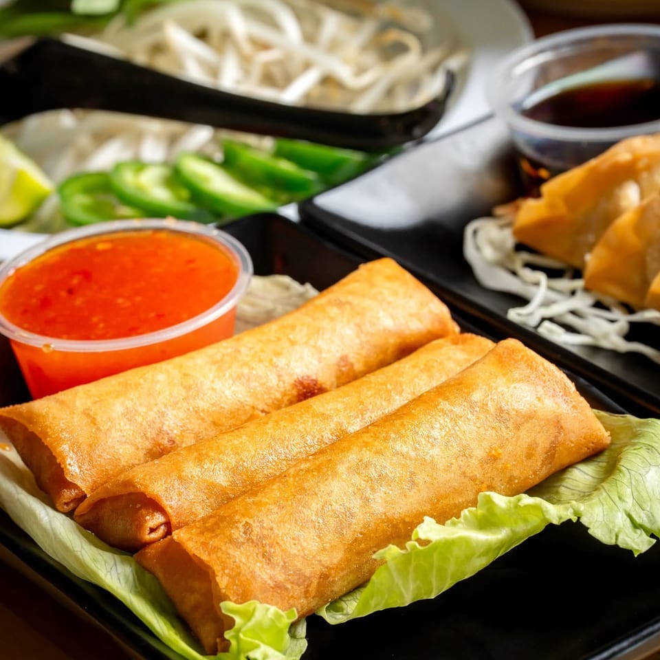 Crispy golden spring rolls served on lettuce with bright sweet chili sauce, noodles, sliced vegetables, and dipping sauce.