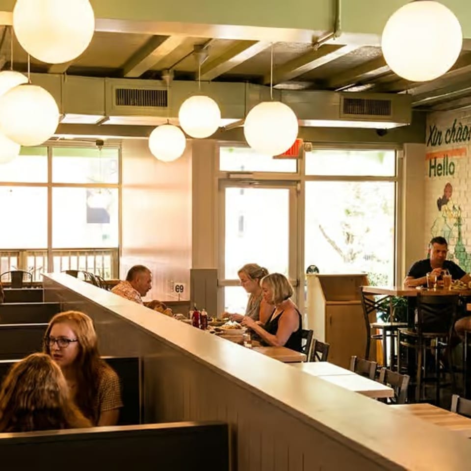 Busy casual restaurant dining room with patrons at tables, globe pendant lights, and colorful mural-covered brick walls.