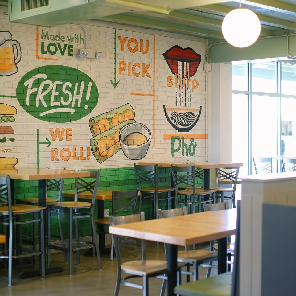Bright restaurant dining area with wooden tables, black chairs, and a colorful mural featuring fresh Vietnamese food and playful lettering.
