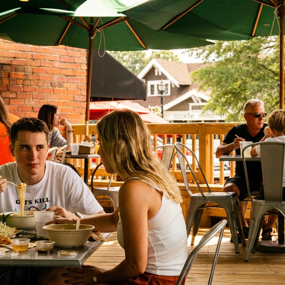 Busy outdoor restaurant patio with diners sharing meals beneath green umbrellas and beside a brick wall.